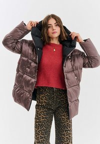 Kurtka puchowa w ciemnym brązie, o błyszczące wykończenie i czarnym kapturze. Teksturowany czerwony sweter pod spodem, w parze z spodniami w wzór panterki.