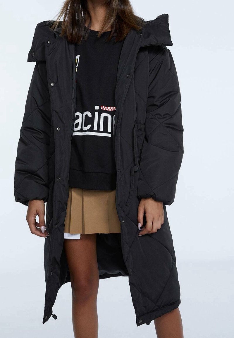 Personne portant un long manteau matelassé noir par-dessus un sweat-shirt graphique noir et une jupe plissée beige, debout devant un fond uni.