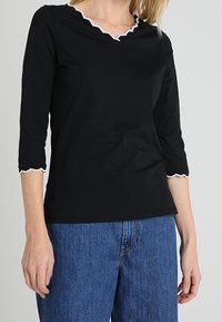 Zwarte t-shirt met een gegolfde halslijn en driekwartmouwen, met witte rand langs de halslijn en mouwopeningen. Casual fit.