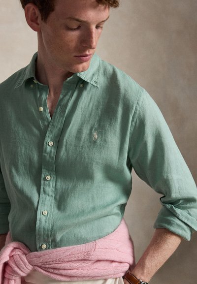 Ung mann som ser ned, iført en grønn knappeskjorte med en liten logo, en rosa genser knyttet rundt livet og en armbåndsur.