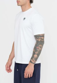 Maglietta bianca a maniche corte in cotone, con scollatura rotonda e un piccolo logo nero sul lato sinistro del petto. Indossata con pantaloni blu con cordoncino.