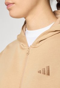Beige vetoketjullinen huppari, joka on valmistettu pehmeästä kankaasta. Siinä on hienovarainen kolmiraidallinen logo, joka on hieman tummemmassa sävyssä, ja teksturoitu pinta.