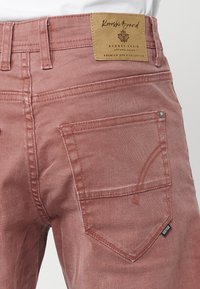Jean en denim rose avec une étiquette en cuir, des détails de couture visibles et une poche accentuée. Présente un design classique à cinq poches.