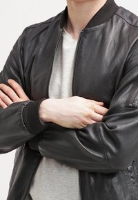Giubbotto bomber in pelle nera con finitura testurizzata, polsini a coste e colletto alto. Chiusura con zip e tasche laterali. Indossato sopra una maglietta grigia.