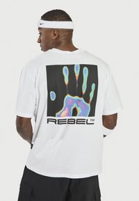 T-shirt en coton blanc avec un graphique carré noir comportant une empreinte colorée et le texte "REBEL™" en dessous. Coupe standard avec des manches courtes.