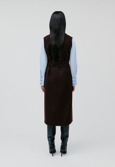 Lange bruine vest met een tailleband, gelaagd over een lichtblauw shirt met knoopdetails, gecombineerd met kniehoge zwarte laarzen.