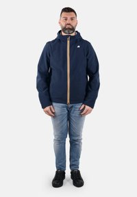 Giacca impermeabile blu navale con zip frontale e accenti arancioni, dotata di cappuccio e maniche aderenti, indossata sopra jeans chiari e scarpe nere.