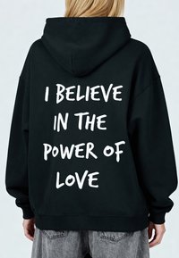 Črni pulover s kapuco z veliko belo napisom na hrbtni strani z besedilom "VERJAMEM V MOČ LJUBEZNI." Ima kapuco z vrvico in rebraste manšete.