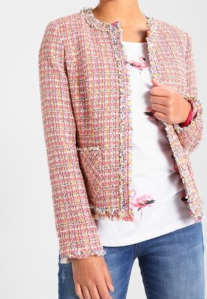 Różowo-biała, teksturowana tweedowa marynarka z frędzlami, noszona na białej koszuli z różowymi nadrukami flamingów, w połączeniu z niebieskimi dżinsami.