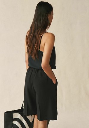 Femeie cu păr blond lung, purtând un top negru fără mâneci și pantaloni scurți negri largi, ținând o geantă tote în dungi alb-negru.