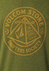 Grøn t-shirt med et gult grafisk design, der viser geometriske former og tekst: "VOLCOM STONE DRIFTERS SOCIETY" og "YOUTH AGAINST ESTABLISHMENT."