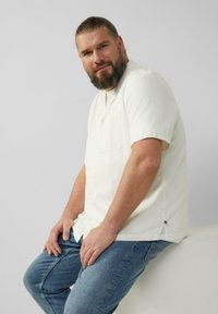 Koszula z krótkim rękawem i kołnierzykiem w jasnym odcieniu kremowym, z teksturowanej tkaniny, z kieszonką z przodu. Model nosi ją z niebieskimi dżinsami i siedzi na białej powierzchni.