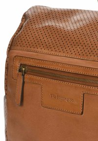 Tan leren tas met een gestructureerd oppervlak en zwarte perforaties. Het heeft een ritsvak en een ingedrukte logo aan de voorkant.