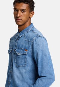 Lichtblauwe denim blouse met een klassieke kraag, twee borstzakken met flappen en goudkleurige knopen. Gestructureerde pasvorm met zichtbare stiksels.