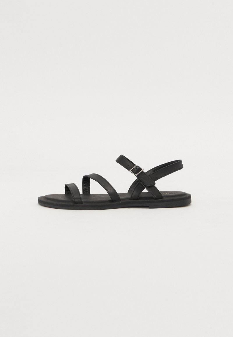 Sandalias de cuero negro con tres correas anchas cruzadas sobre el pie y una correa ajustable en el tobillo. Suela plana con un mínimo de dibujo. Diseño simple.