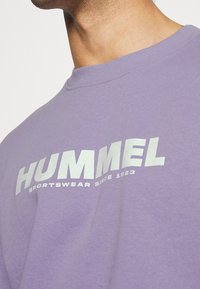 Levandulové bavlněné tričko s kulatým výstřihem a krátkými rukávy. Na přední straně vynikající bílý text "HUMMEL SPORTSWEAR SINCE 1923".
