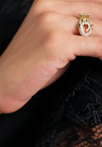 Zlatý hadí prsteň so vzorom s bielymi kamenčekmi, červeným detailom a tmavomodrými drahokamami. Ruka s prstenom na čiernom čipkovanom pozadí.