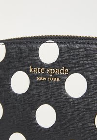 Portefeuille noir en texture avec des pois blancs, arborant un logo doré "kate spade NEW YORK" à l'avant et une fermeture éclair dorée.