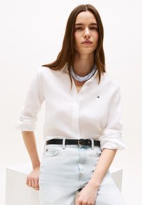 Ung kvinne iført hvit knyteskjorte, lyseblå jeans med svart belte og blåstripet halseskjerf, poserer foran en ensfarget bakgrunn.