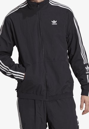 Mężczyzna ubrany w czarny dres Adidas z białymi paskami na rękawach i spodniach, lewa ręka w kieszeni, stojący na białym tle.