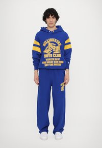 Blaues Hoodie- und Sweatpants-Set mit gelbem Grafiktext und Akzenten. Verfügt über eine lockere Passform, Baumwollmaterial und weiße Sneakers.
