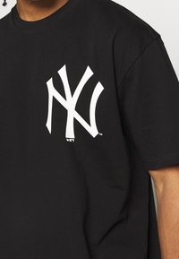 Svart T-shirt med en stor vit New York Yankees-logotyp på vänster bröst, bärs av en person med medelhudton.