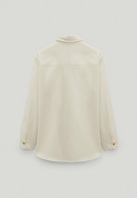 Chemise beige à manches longues avec col, poignets boutonnés et couture droite de l'empiècement dos, présentée à plat sur un fond clair.