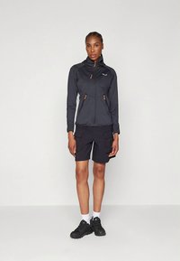 Lengvas juodas užtrauktuku užsegamas švarkelis su oranžiniais akcentais, derinamas su juodais krovinių šortais. Modelis vilki juodus sportinius batelius ir baltas kojines.