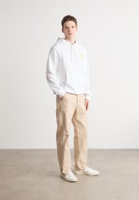 Felpa bianca con logo "HUGO" giallo, pantaloni cargo beige e sneakers bianche. Outfit casual con maniche lunghe e tasca frontale a marsupio.