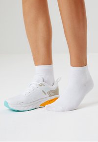 Vit sportskor med turkosa och orange detaljer, texturerade meshnätpaneler och snörningssystem. Matchas med en vit ankelsocka på den andra foten.