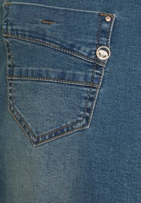 Denimkangas, jossa on haalistunut sininen viimeistely, takataskussa on oranssit tikit ja metallinen logo lähellä taskun ykköstä.
