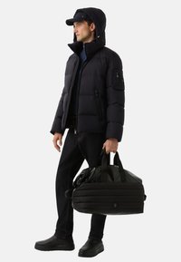 Pufferjack in zwart met een capuchon, met ritszakken en een gladde textuur, gecombineerd met donkere jeans en een zwarte sporttas.