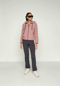 Rosa fleece-jakke med hette, glidelås foran og to sidelommer, kombinert med mørke bukser og beige sneakers. Solbriller brukt.