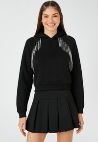 Sudadera negra con detalles de flecos plateados en la parte frontal, puños y dobladillo acanalados; combinada con una falda corta plisada negra. Textura de tela suave.