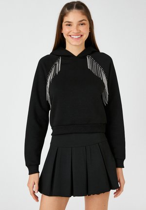 Sudadera negra con detalles de flecos plateados en la parte frontal, puños y dobladillo acanalados; combinada con una falda corta plisada negra. Textura de tela suave.
