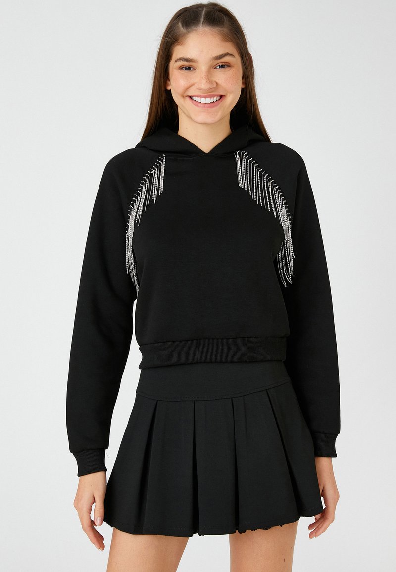Sudadera negra con detalles de flecos plateados en la parte frontal, puños y dobladillo acanalados; combinada con una falda corta plisada negra. Textura de tela suave.