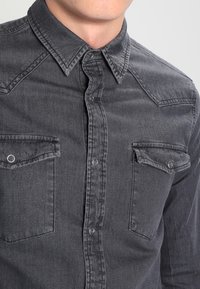 Man som bär en urtvättad svart denimskjorta med spetsig krage och två knäppta bröstfickor.