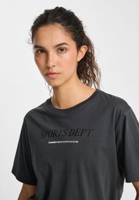 Svart bomullströja med korta ärmar, med "SPORTS DEPT." och "hummel PERFORMANCE" logotyper i kontrasterande färger på framsidan.