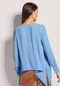 Hellblaues Langarmbluse mit fließendem Design, seitlichen Schlitzen und geknöpften Manschetten. Glatter Stoff mit lockerer Passform.