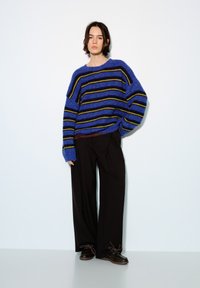 Pull en maille oversize à rayures bleues et noires avec une coupe décontractée, associé à un pantalon large noir et des chaussures à lacets marron.
