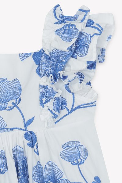 Λευκό φόρεμα από ύφασμα με μπλε κεντήματα λουλουδιών, λεπτομέρεια σούρας στους ώμους και ζώνη στη μέση. Διαθέτ ει περίπλοκα μοτίβα πετάλων και υφή.