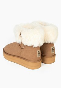 Botas de tobillo de ante marrón con un borde de piel sintética blanca. Cuentan con una suela de goma plana en tono beige y un pequeño parche con el logo en el talón.
