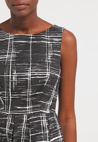 Sort og hvid ærmeløs kjole med et struktureret mønster af krydsende linjer, tætsiddende liv og plisseret skørt.