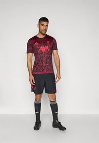 Maillot de sport rouge à motifs avec manches courtes, comprenant des accents noirs et un logo, associé à un short noir et des chaussettes montantes noires.