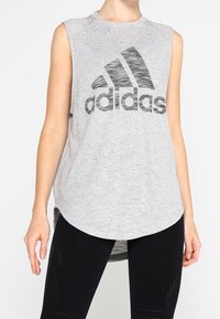Harmaa hihaton urheiluostos, joka on valmistettu teksturoidusta kankaasta ja jossa on suuri musta Adidas-logo edessä, hieman kaarevalla helmaksi.
