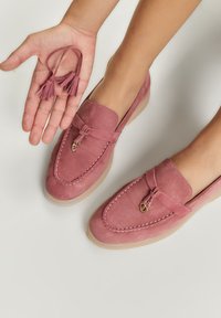 Due mocassini in suede rosa con un design cucito, caratterizzati da un decorativo fiocco e hardware dorato. Una mano tiene delle frange rosa abbinate.