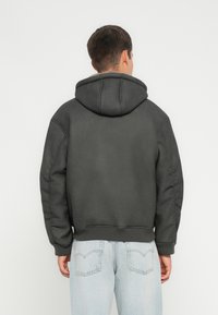 Sweat à capuche gris foncé avec une coupe décontractée, des poignets et un ourlet côtelés, et un tissu texturé. Porté avec un jean en denim bleu clair.