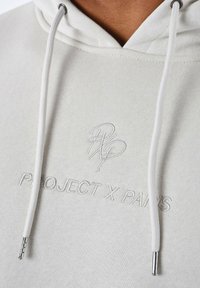 Sudadera gris claro con cordones, que presenta el logo bordado "PROJECT X PARIS". Textura suave, grosor medio, detalles metálicos en los cordones.