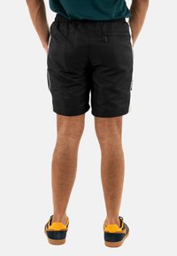 Bañador negro con cintura elástica, rayas blancas a los lados y un bolsillo trasero. Complementado con zapatillas amarillas y marinas.