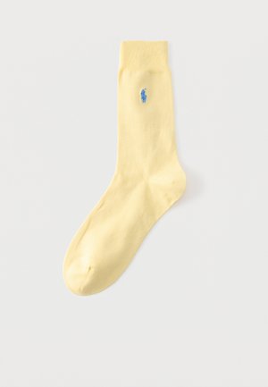 Ein einzelner hellgelber Socken mit geripptem Bund und kleinem blau besticktem Polo-Spieler-Logo nahe dem oberen Rand vor einfarbigem Hintergrund.